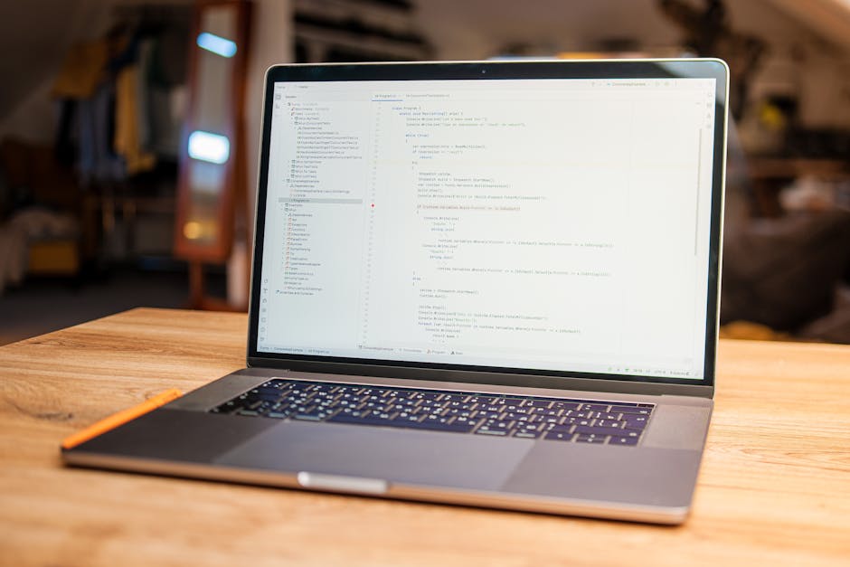 This image features a close-up view of a silver laptop placed on a wooden desk with a smooth surface, displaying lines of code or text on its screen. The background is softly blurred, suggesting an in